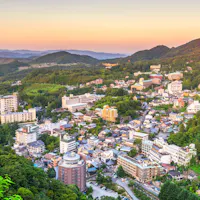 Arima Onsen Aerial view of a town nestled among green mountains at sunset, with numerous buildings and hotels scattered across the hilly landscape under a colorful sky.