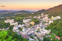 Aerial view of a town nestled among green mountains at sunset, with numerous buildings and hotels scattered across the hilly landscape under a colorful sky.