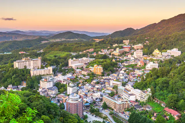 Aerial view of a town nestled among green mountains at sunset, with numerous buildings and hotels scattered across the hilly landscape under a colorful sky.