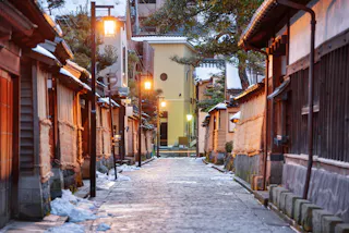 A quiet, narrow stone street lined with traditional Japanese wooden houses, softly lit by warm lanterns in the early evening. Patches of snow are visible on the ground and rooftops.