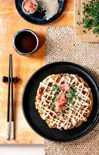 Okonomiyaki A plate of okonomiyaki topped with mayonnaise, sauce, chopped chives, pickled ginger, and sesame seeds, with chopsticks, a dipping sauce, and chopped chives on the side.