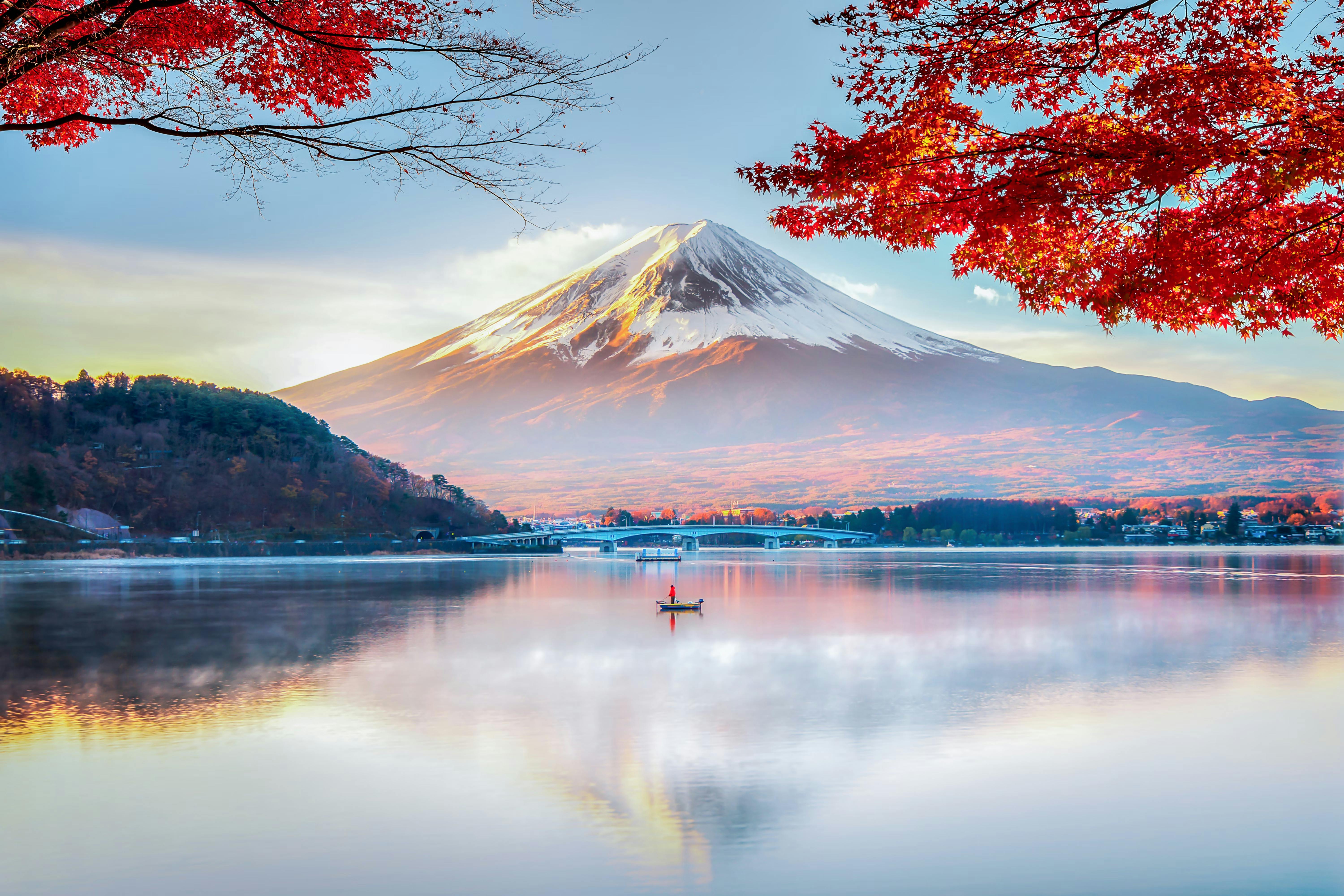 A person in a red kayak paddles on a calm lake, with Mount Fuji in the background and vibrant red autumn leaves framing the scene.
