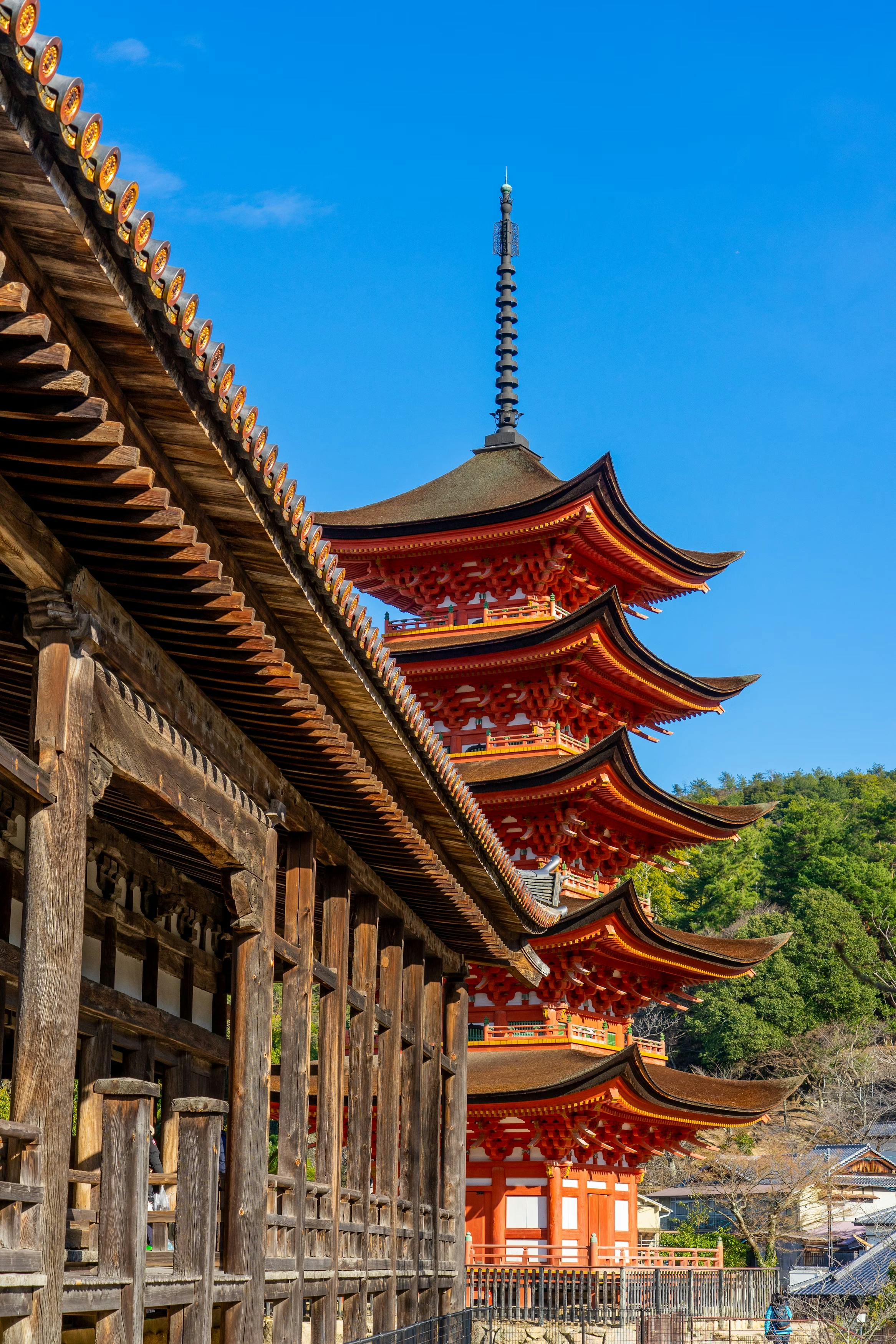 A bright red five-story pagoda stands beside a traditional wooden building under a clear blue sky, surrounded by green trees and hills.