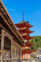 A bright red five-story pagoda stands beside a traditional wooden building under a clear blue sky, surrounded by green trees and hills.