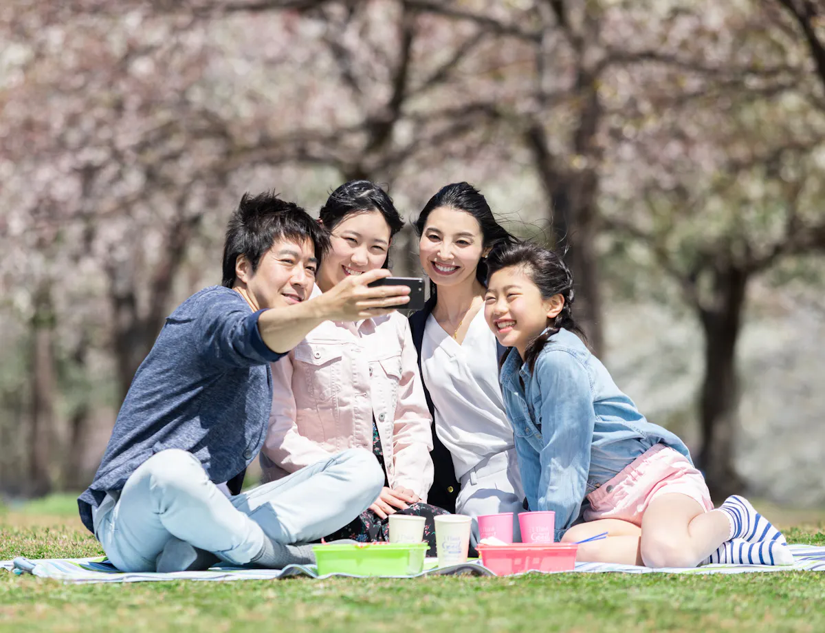 Hanami Picnic A family of four sits on a picnic blanket outdoors, smiling and taking a selfie together. Cherry blossom trees bloom in the background, and picnic items are arranged on the blanket in front of them.