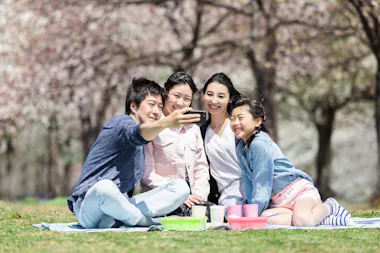 A family of four sits on a picnic blanket outdoors, smiling and taking a selfie together. Cherry blossom trees bloom in the background, and picnic items are arranged on the blanket in front of them.