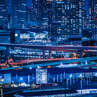 Odaiba A vibrant cityscape at night with tall illuminated buildings, elevated highways, and bright city lights. The image shows urban infrastructure, glowing windows, and busy roads, creating a lively metropolitan atmosphere.