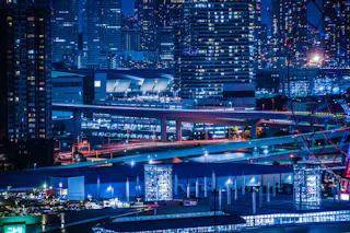 A vibrant cityscape at night with tall illuminated buildings, elevated highways, and bright city lights. The image shows urban infrastructure, glowing windows, and busy roads, creating a lively metropolitan atmosphere.