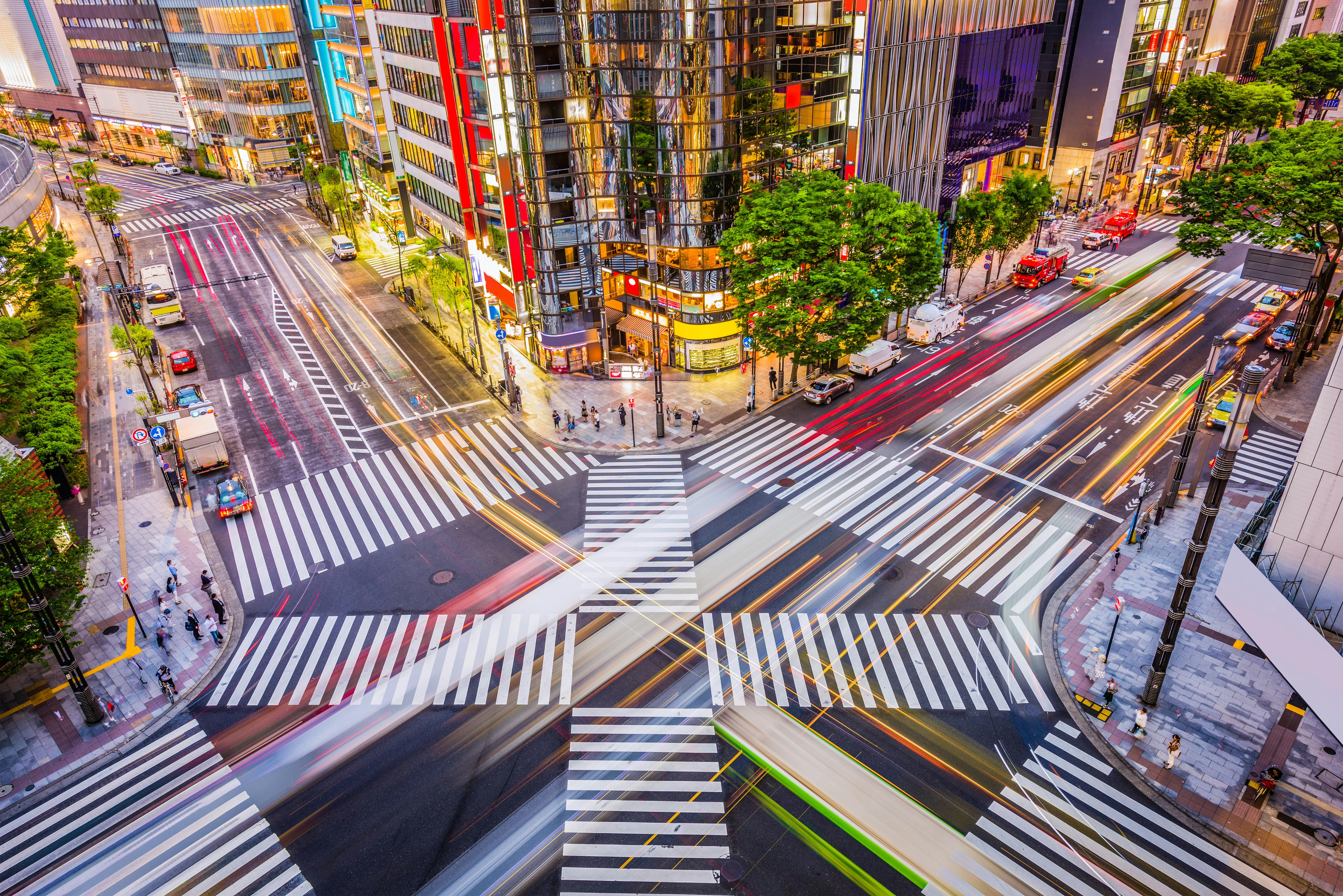 Aerial view of a busy city intersection at night with crosswalks, blurred car lights, tall modern buildings, and some trees lining the streets. Pedestrians are crossing and traffic flows in multiple directions.