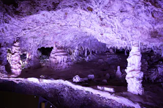 Illuminated cave interior featuring jagged stalactites hanging from the ceiling and stalagmites rising from the floor, all bathed in purple-tinted light.