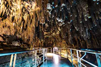 A metal walkway with railings leads through a cave filled with jagged stalactites hanging from the ceiling, illuminated by artificial lights.