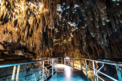 A metal walkway with railings leads through a cave filled with jagged stalactites hanging from the ceiling, illuminated by artificial lights.