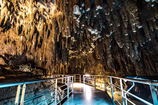 A metal walkway with railings leads through a cave filled with jagged stalactites hanging from the ceiling, illuminated by artificial lights.