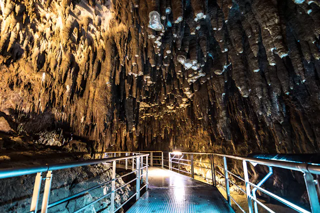 A metal walkway with railings leads through a cave filled with jagged stalactites hanging from the ceiling, illuminated by artificial lights.