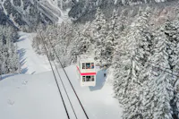 An aerial cable car travels over a snowy mountain landscape, surrounded by pine trees covered in snow, with distant peaks in the background.