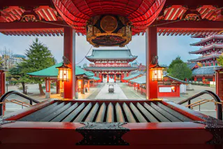 View from inside a traditional Japanese temple, featuring red pillars, lanterns, and a main building in the background. The scene includes a courtyard and green-roofed structures, likely at Senso-ji Temple in Tokyo.