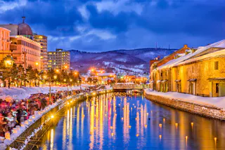 A snowy canal lined with old warehouses, glowing streetlights, and reflections in the water at dusk, with mountains and modern buildings in the background under a blue, cloudy sky.