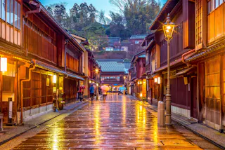 A narrow, rain-soaked street lined with traditional wooden buildings and glowing lanterns in the evening, with blurred figures walking and green trees visible in the background.