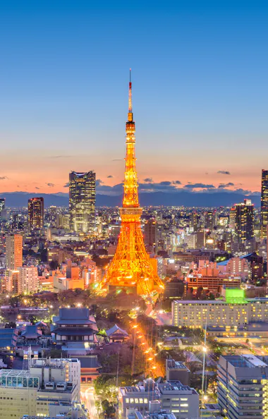 Tokyo Tokyo cityscape at dusk with Tokyo Tower illuminated in orange at the center, skyscrapers surrounding it, and Mount Fuji visible in the distant background under a clear sky.