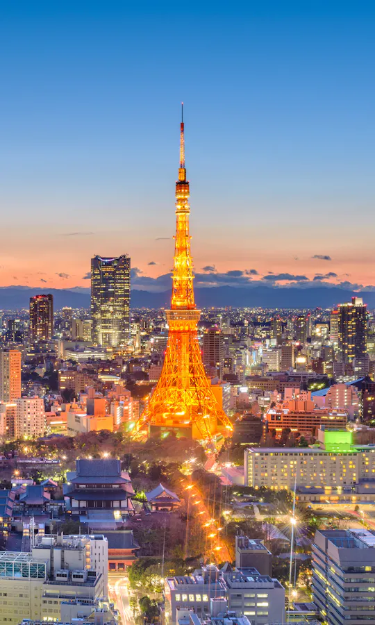 Tokyo Tokyo cityscape at dusk with Tokyo Tower illuminated in orange at the center, skyscrapers surrounding it, and Mount Fuji visible in the distant background under a clear sky.