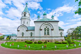 A white church with green domes and arched windows stands on a grassy lawn with small shrubs and a curved pathway, under a bright blue sky with scattered clouds.