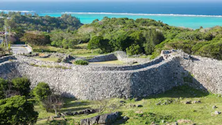 Ancient stone walls of a historic castle ruin surrounded by lush greenery, with turquoise ocean water and blue sky in the background.
