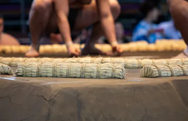 Close-up of a sumo wrestling ring made of clay with straw bales marking the boundary, with two sumo wrestlers crouched and preparing to engage in the background.