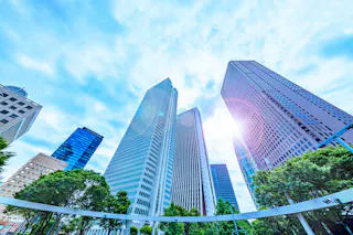 Skyscrapers viewed from below against a bright blue sky with scattered clouds. Sunlight shines through the buildings, and green trees are visible at the base of the towers.