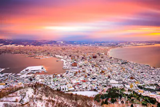 A vibrant sunset sky casts colorful light over a coastal city surrounded by mountains and snowy landscapes, with buildings clustered along the shoreline and two bays.