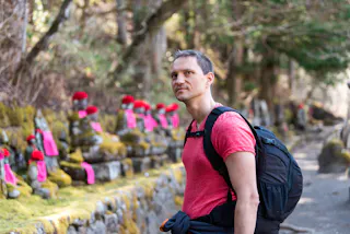 A man with a backpack stands on a stone path lined with moss-covered statues wearing red hats and bibs, surrounded by trees in a peaceful outdoor setting.
