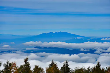 A distant mountain range is seen under a blue sky, with layers of clouds and mist partially covering the landscape. Tall trees with autumn foliage are visible in the foreground.