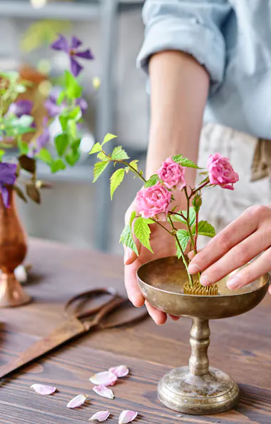 Ikebana Japan flower arrangement A person arranges small pink flowers in a metal vase on a wooden table, surrounded by gardening tools, flower petals, and another vase with purple flowers.