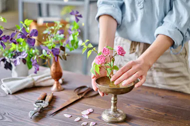 A person arranges small pink flowers in a metal vase on a wooden table, surrounded by gardening tools, flower petals, and another vase with purple flowers.