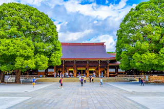 People walk and gather in front of a large traditional Japanese shrine with a wide, sloped roof, flanked by two lush green trees, under a bright blue sky with scattered clouds.