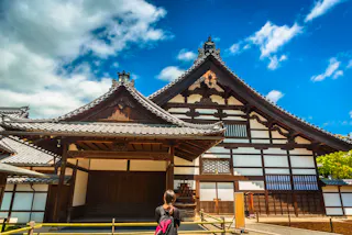 A person with a backpack stands facing a traditional Japanese building with ornate wooden roofs and white walls, under a bright blue sky with scattered clouds.