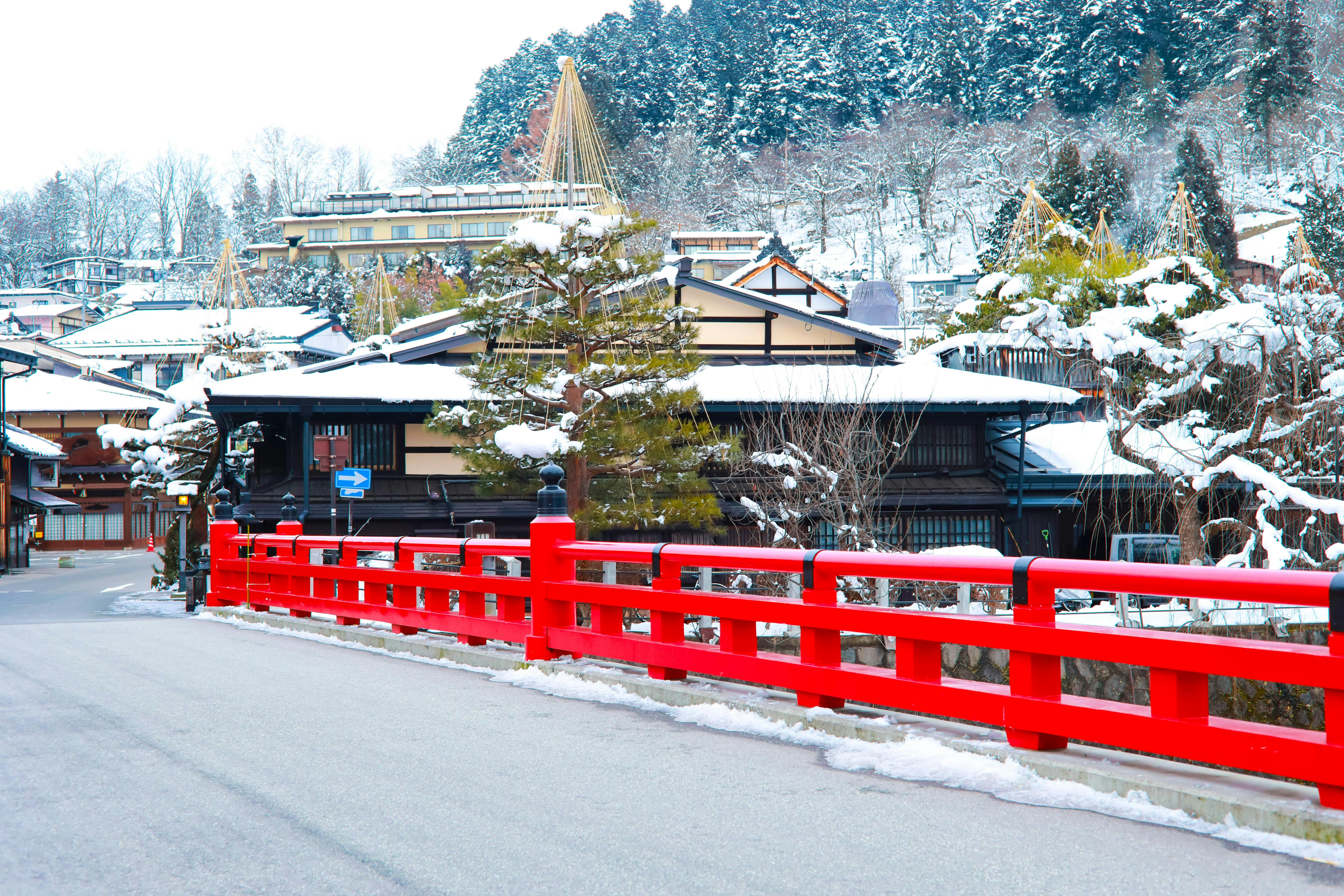 A bright red bridge crosses over a snowy street in front of traditional Japanese buildings and pine trees, with snow-covered rooftops and a forested hillside in the background.