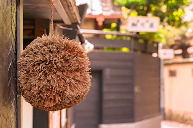 A large, round ball made of brown cedar branches hangs outside a building in a sunlit, traditional Japanese street. The background is softly blurred, showing wooden structures and greenery.