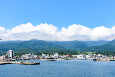 A coastal town with white buildings sits by the water, backed by lush green mountains under a blue sky with clouds. Boats are docked at the harbor in the foreground.