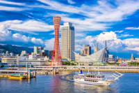 A white sightseeing boat sails in a harbor with modern buildings, a red observation tower, and a unique white structure in the background under a bright blue sky with clouds.