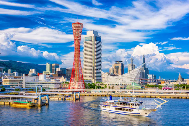 A white sightseeing boat sails in a harbor with modern buildings, a red observation tower, and a unique white structure in the background under a bright blue sky with clouds.