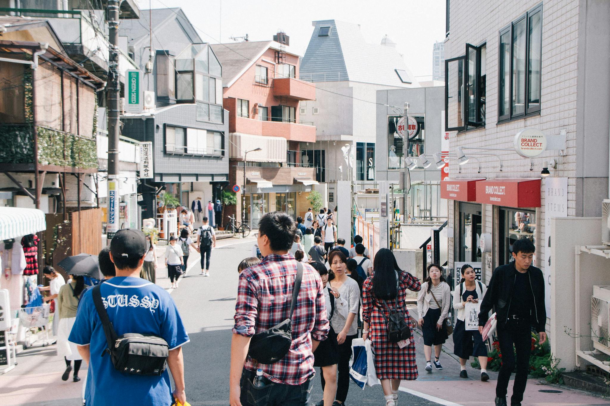 A busy street scene in a Japanese city with people walking, shopping, and talking. Buildings with shops, balconies, and signs line both sides of the street under a bright sky.