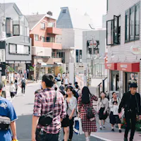 Cat Street Shibuya A busy street scene in a Japanese city with people walking, shopping, and talking. Buildings with shops, balconies, and signs line both sides of the street under a bright sky.