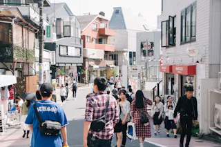 A busy street scene in a Japanese city with people walking, shopping, and talking. Buildings with shops, balconies, and signs line both sides of the street under a bright sky.