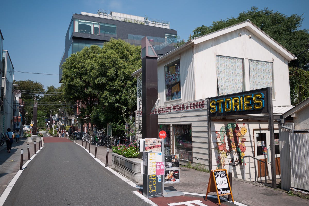 A narrow street lined with trees and bicycles features a white building with a neon “STORIES” sign, colorful posters, and sale signs outside. Modern glass buildings and other shops are visible in the background on a sunny day.