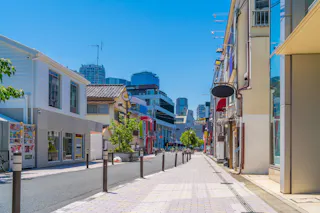 A quiet city street lined with modern and traditional buildings, trees, and storefronts under a clear blue sky, with tall skyscrapers visible in the background.