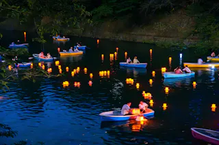 People in rowboats float on a river at night, surrounded by glowing lanterns on the water. Trees and an embankment frame the scene, creating a peaceful, festive atmosphere.