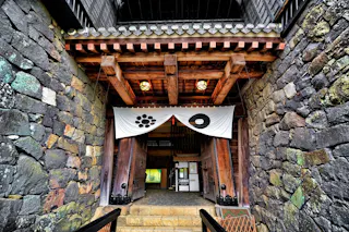 Entrance of a traditional Japanese castle with large wooden doors, stone walls, and white banners featuring black symbols hanging above the doorway. Warm lights illuminate the wooden beams and interior.