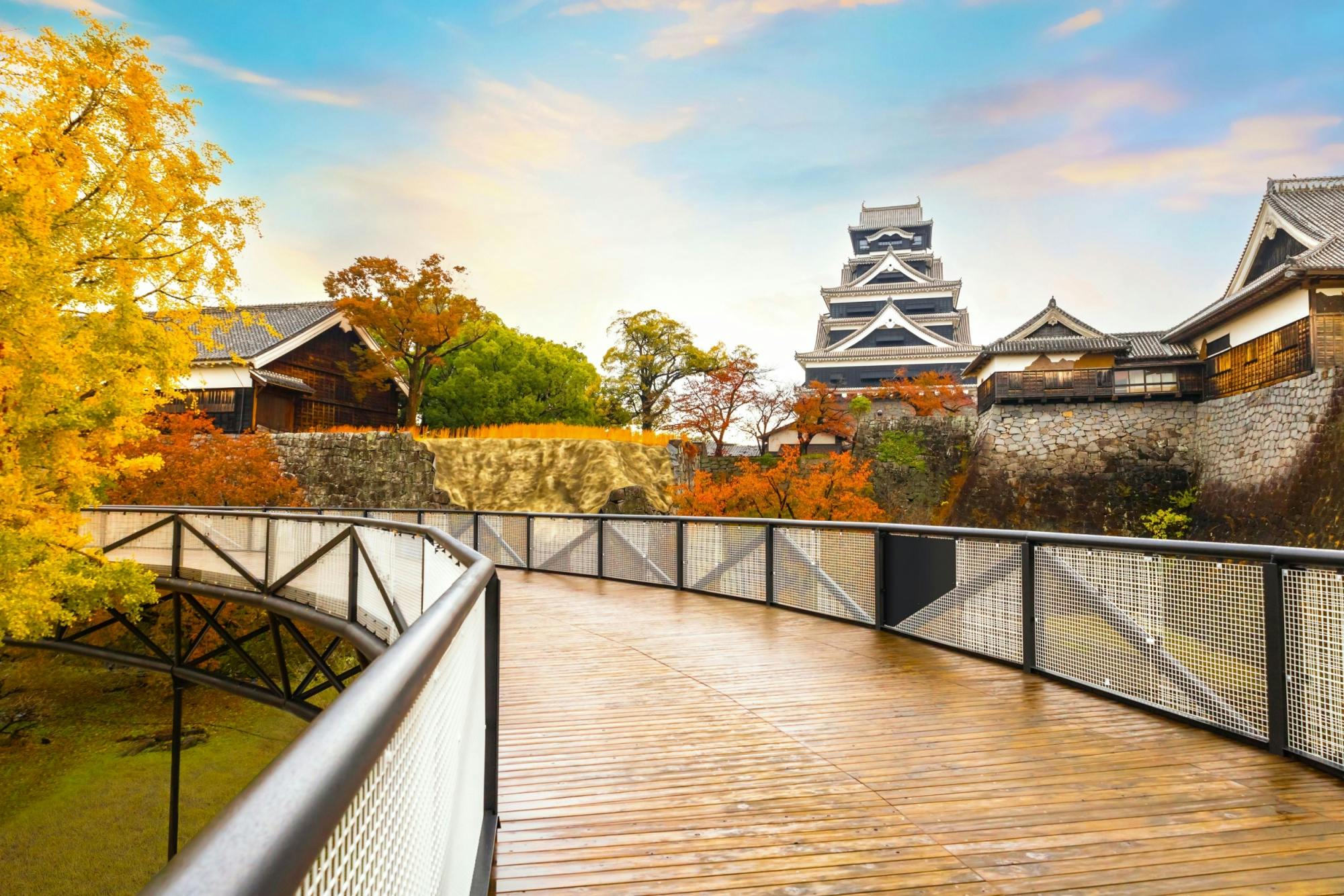 A wooden walkway leads to a traditional Japanese castle surrounded by stone walls and buildings, with autumn trees and a bright, colorful sky in the background.