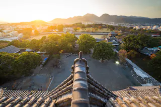 View from a high point of a traditional Japanese castle roof overlooking a courtyard, trees, distant city buildings, and mountains at sunset, with warm sunlight illuminating the scene.