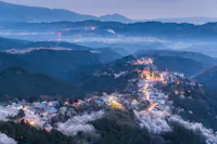 Aerial view of a mountain village at dusk, with illuminated streets winding among cherry blossom trees, surrounded by forested hills and mist, and distant mountains in the background.